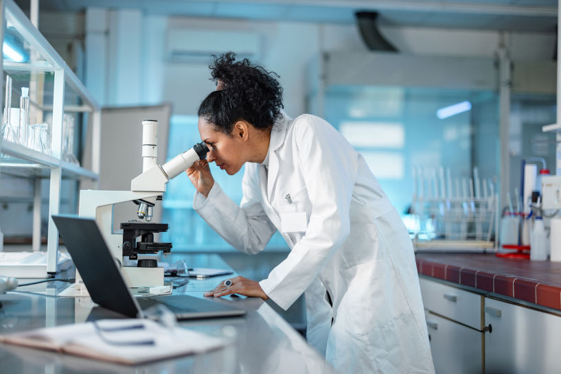 Hispanic scientist in lab coat, studying under microscope and using laptop in a lab